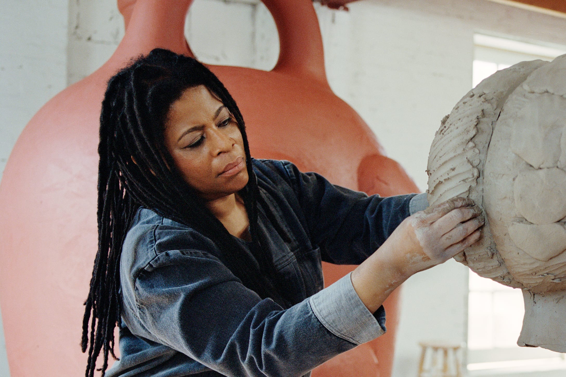 Simone Leigh, a medium-dark-skinned woman with dreadlocks working on a large clay sculpture in a denim work outfit